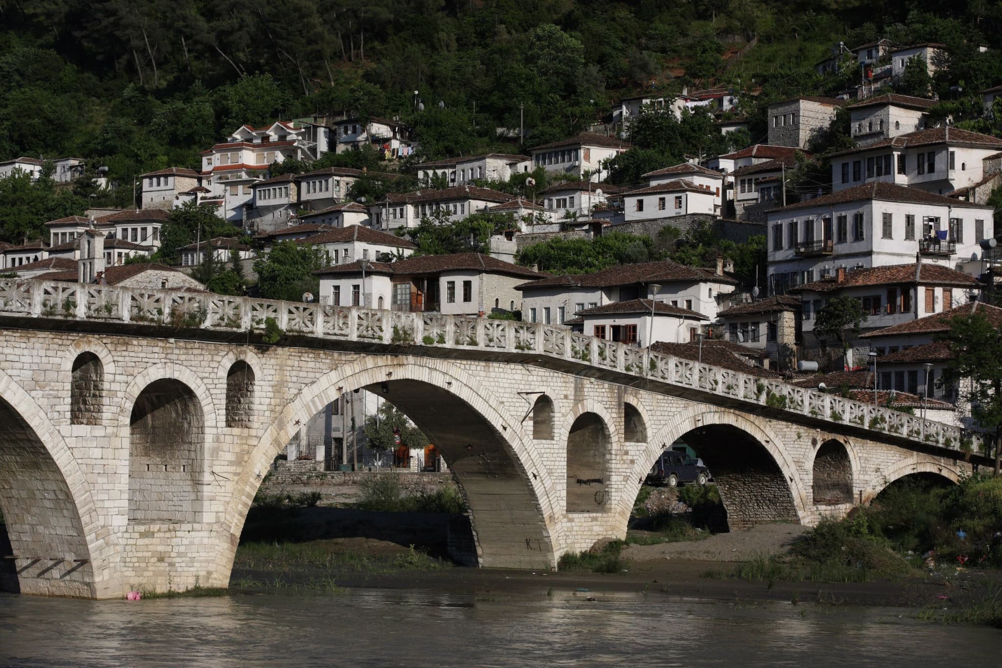 Berat in Albanië: een oude stenen brug met bogen over de rivier de Osum, en daarachter witte huizen met rode daken tegen een groene heuvel.