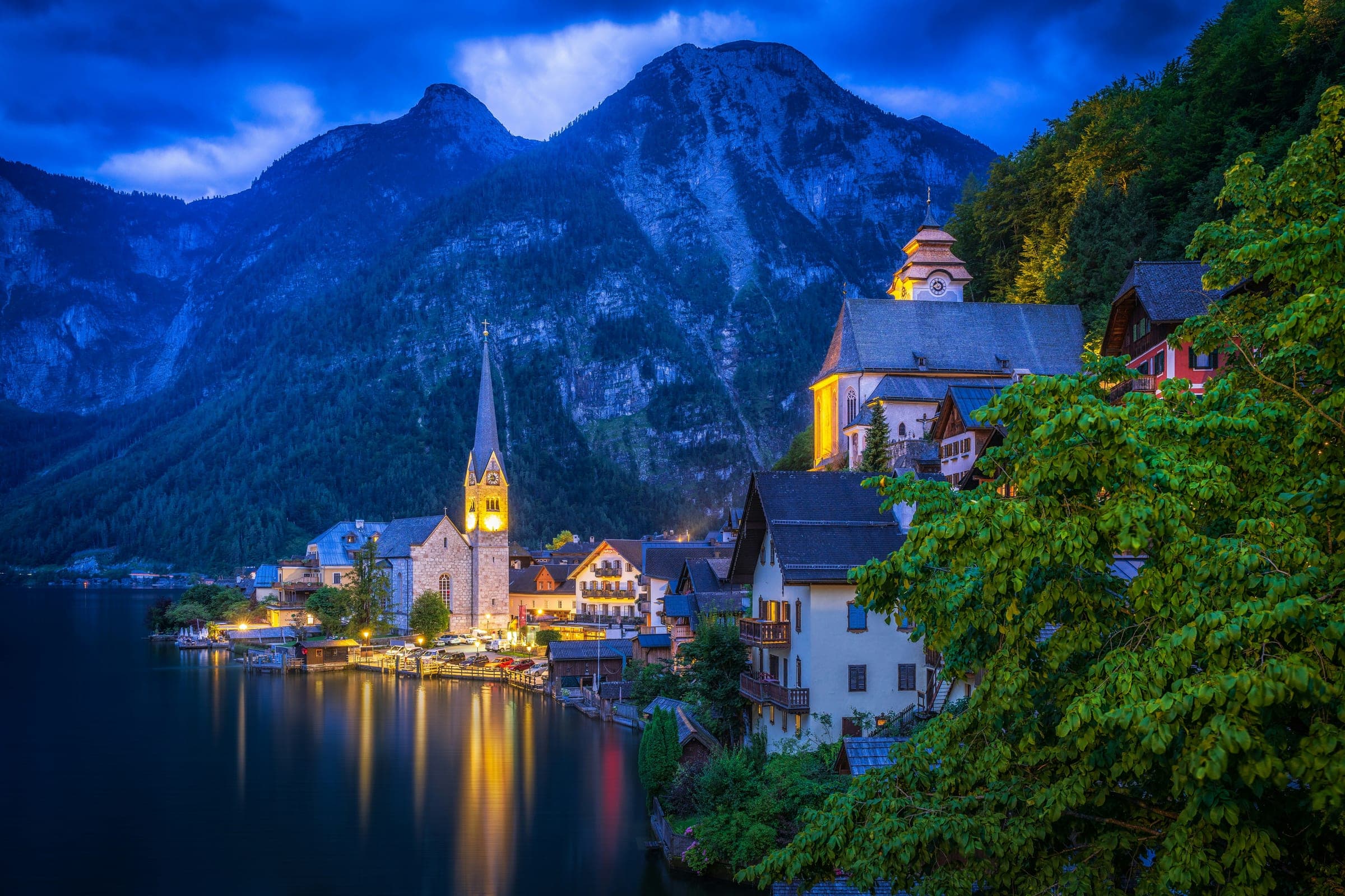 Hallstatt in het blauwe uur: het verlichte dorp met de kerktoren aan het stille meer, de bergen donker erboven.
