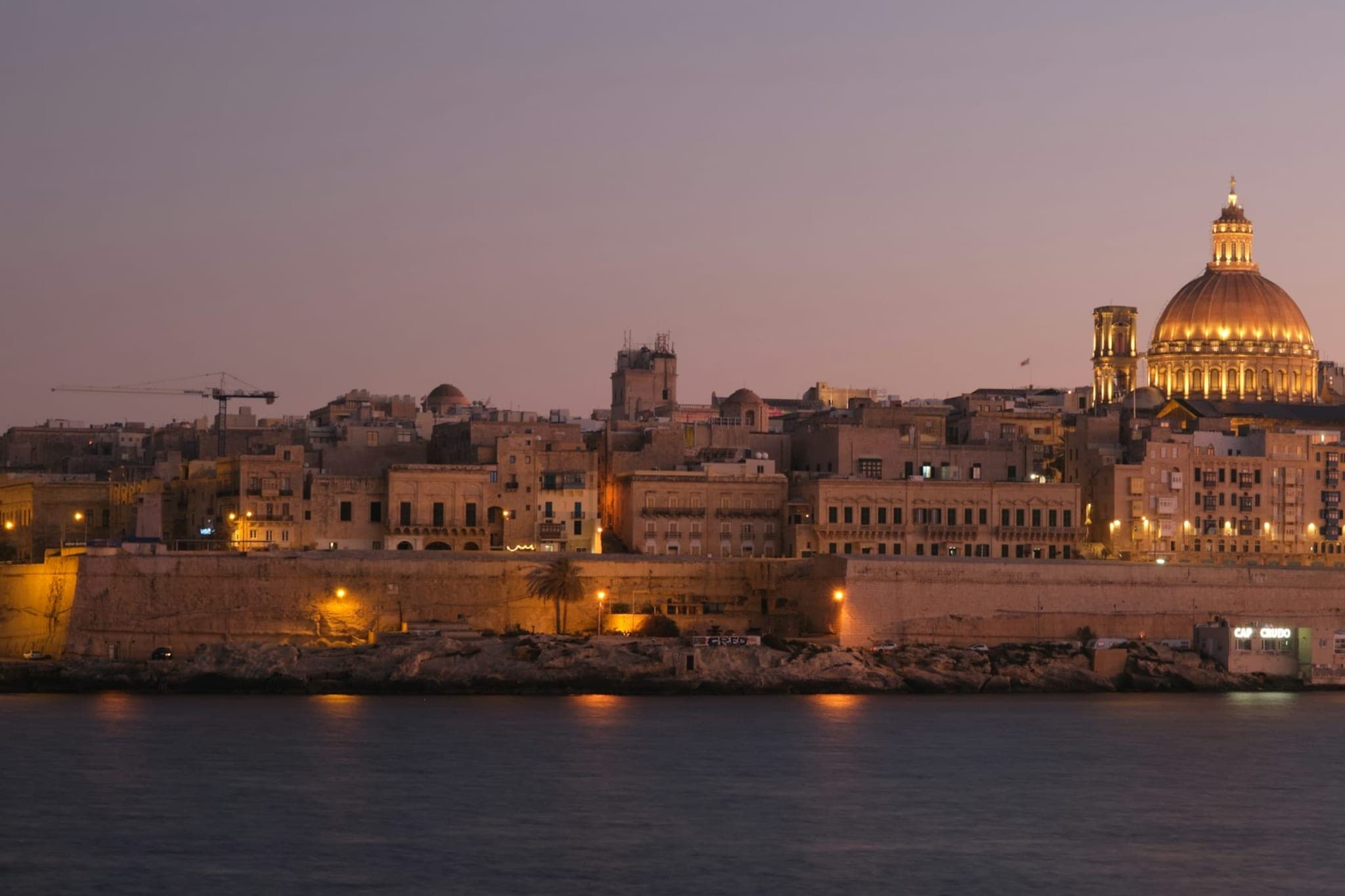 Valletta at dusk, seen across the water, with the lit dome of the Carmelite church above the fortress wall.