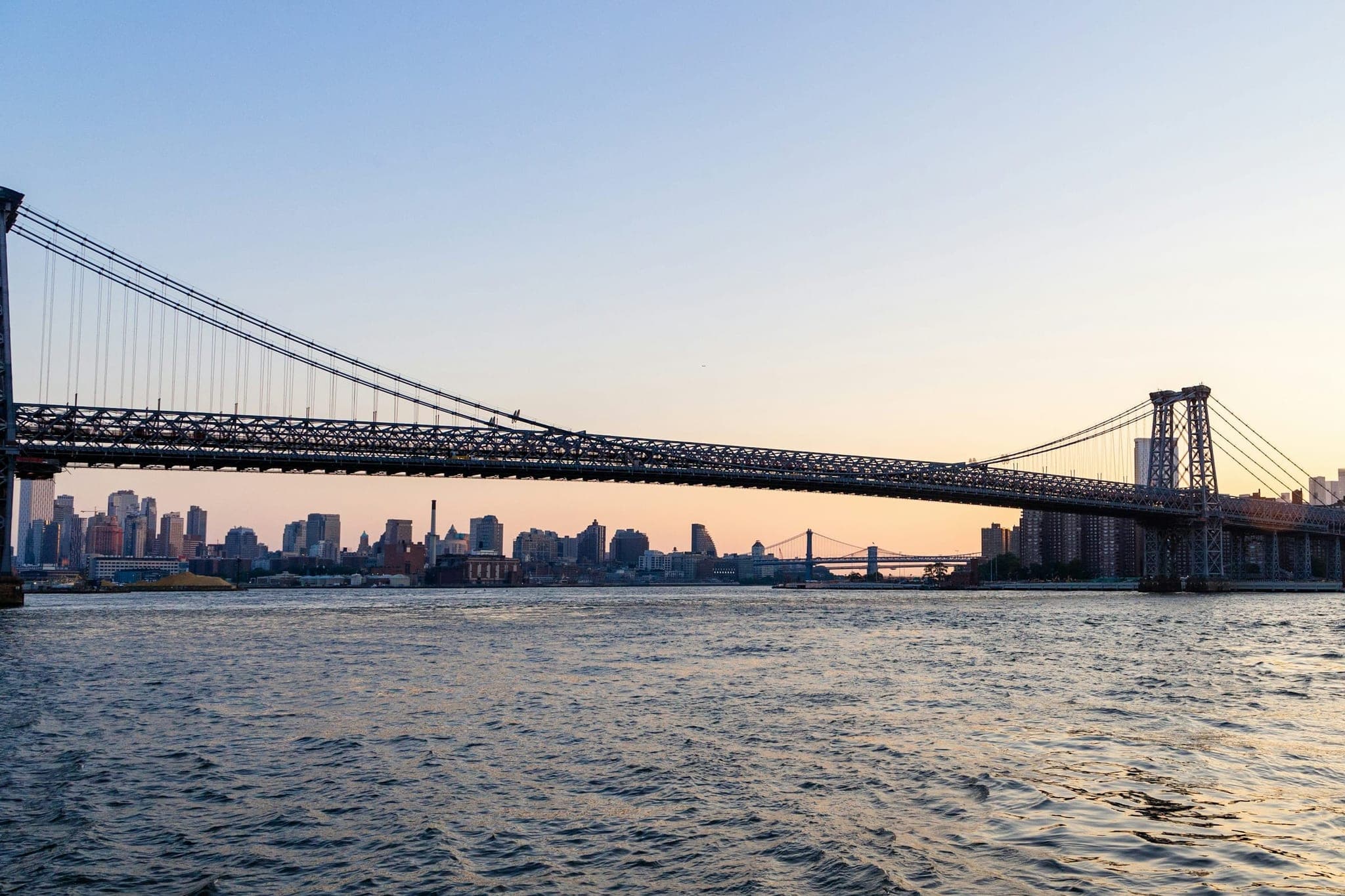 The Williamsburg Bridge over the East River at dusk, with the New York skyline behind it.