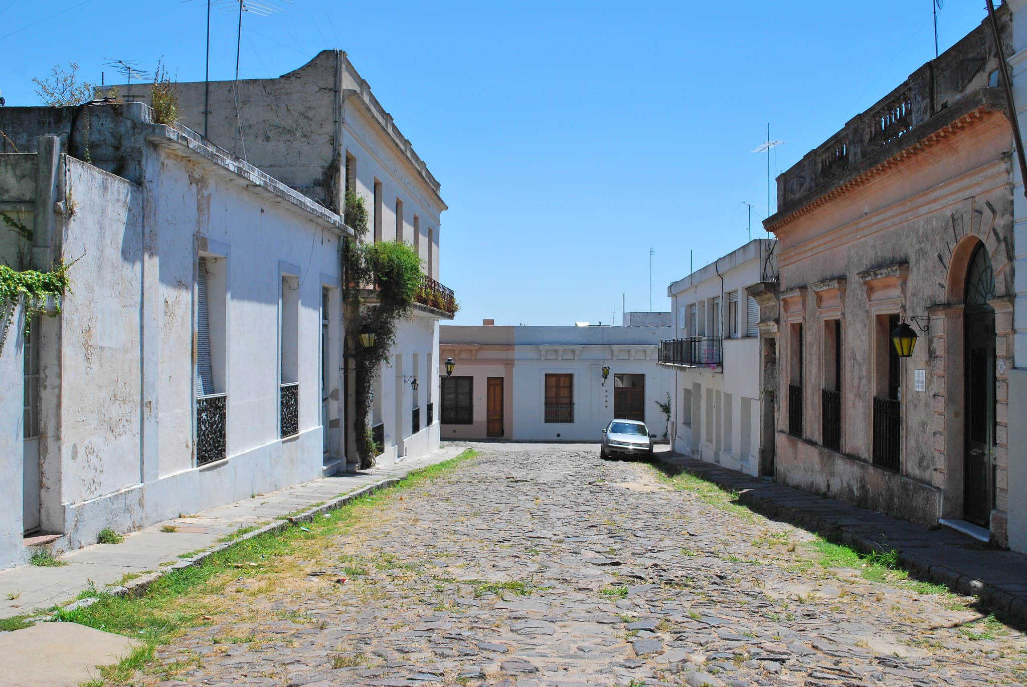 Een oude keienstraat in Colonia del Sacramento: lage witte koloniale huizen met smeedijzeren ramen en lantaarns, onder een strakblauwe lucht.