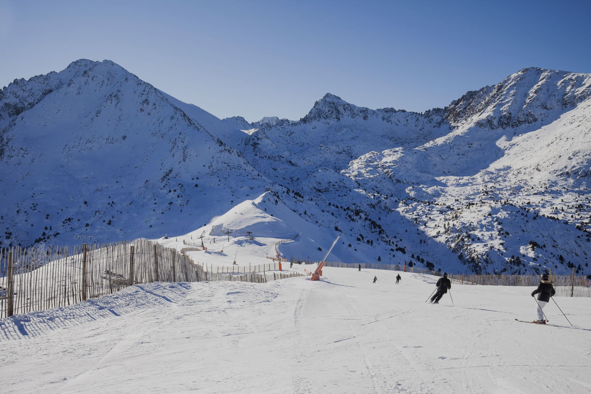 Een skipiste in Grandvalira in Andorra op een heldere winterdag: besneeuwde bergtoppen, een hek langs de piste en twee skiërs in de zon.