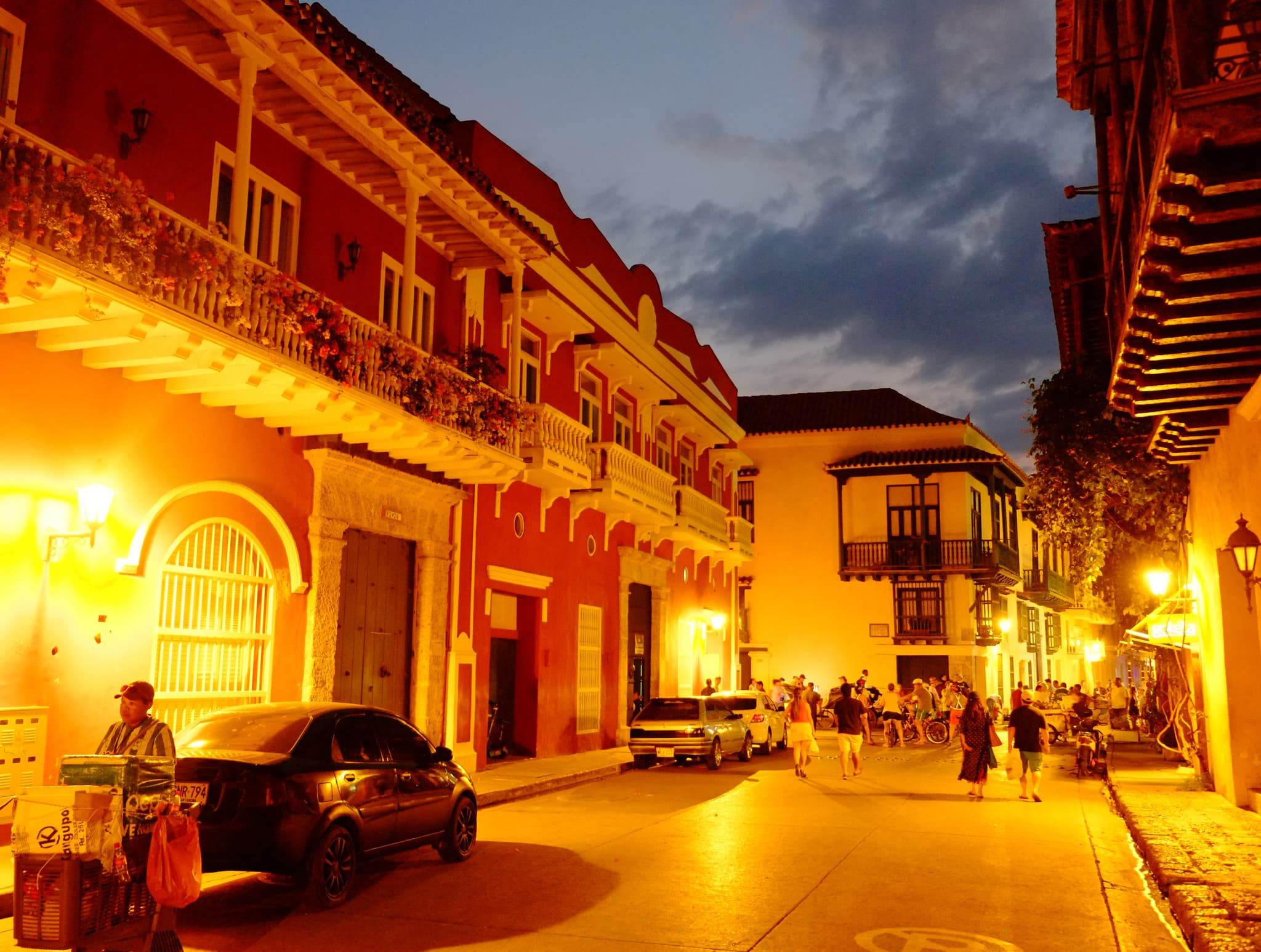 A street in the walled city of Cartagena at dusk: red and yellow colonial houses with wooden balconies full of flowers, lit by street lamps, with people strolling at the corner.