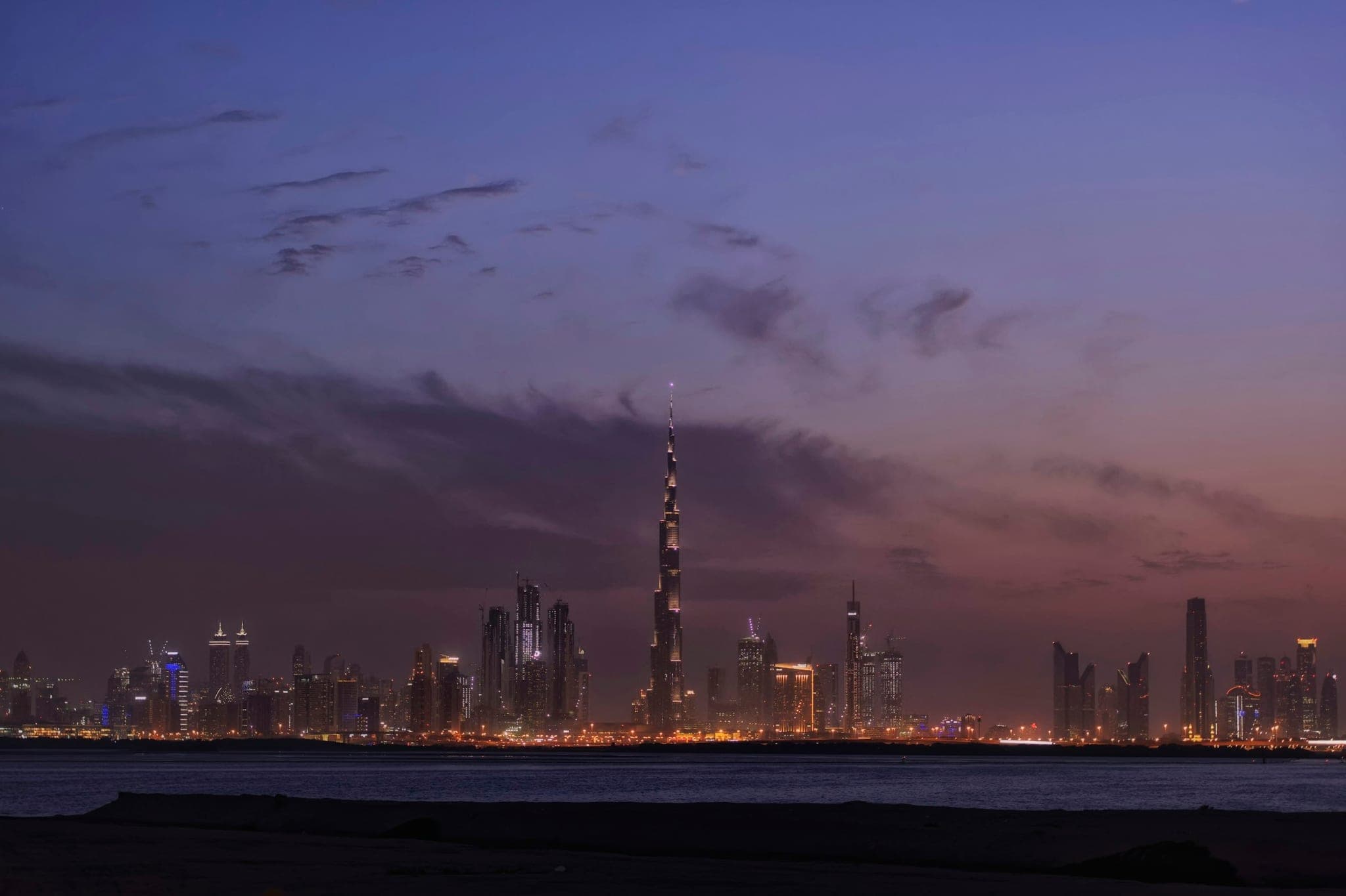 The Dubai skyline in the evening, seen across the water, with the lit towers reflected in the surface.