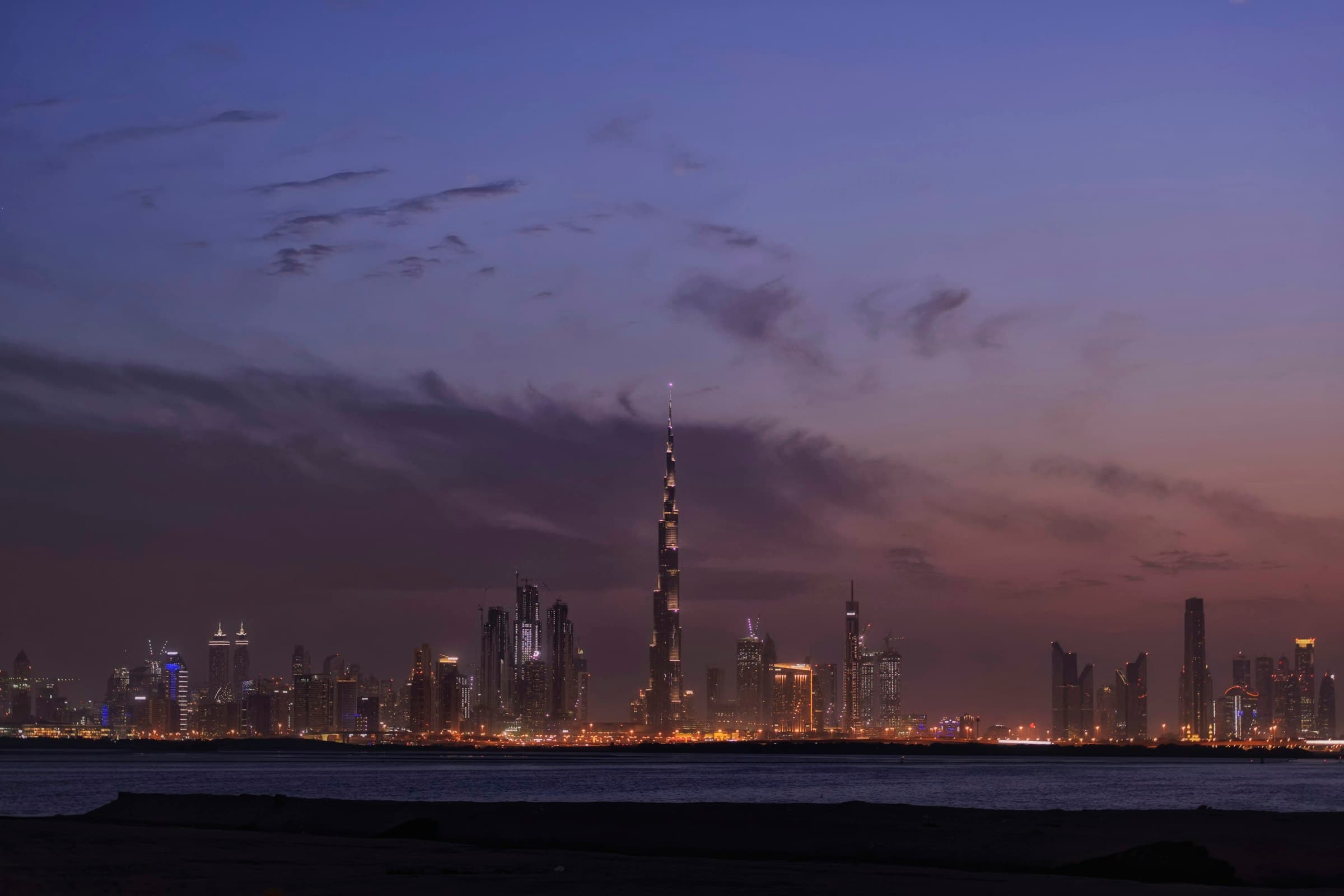 De skyline van Dubai in de avond, gezien over het water, met de verlichte torens weerspiegeld in het wateroppervlak.