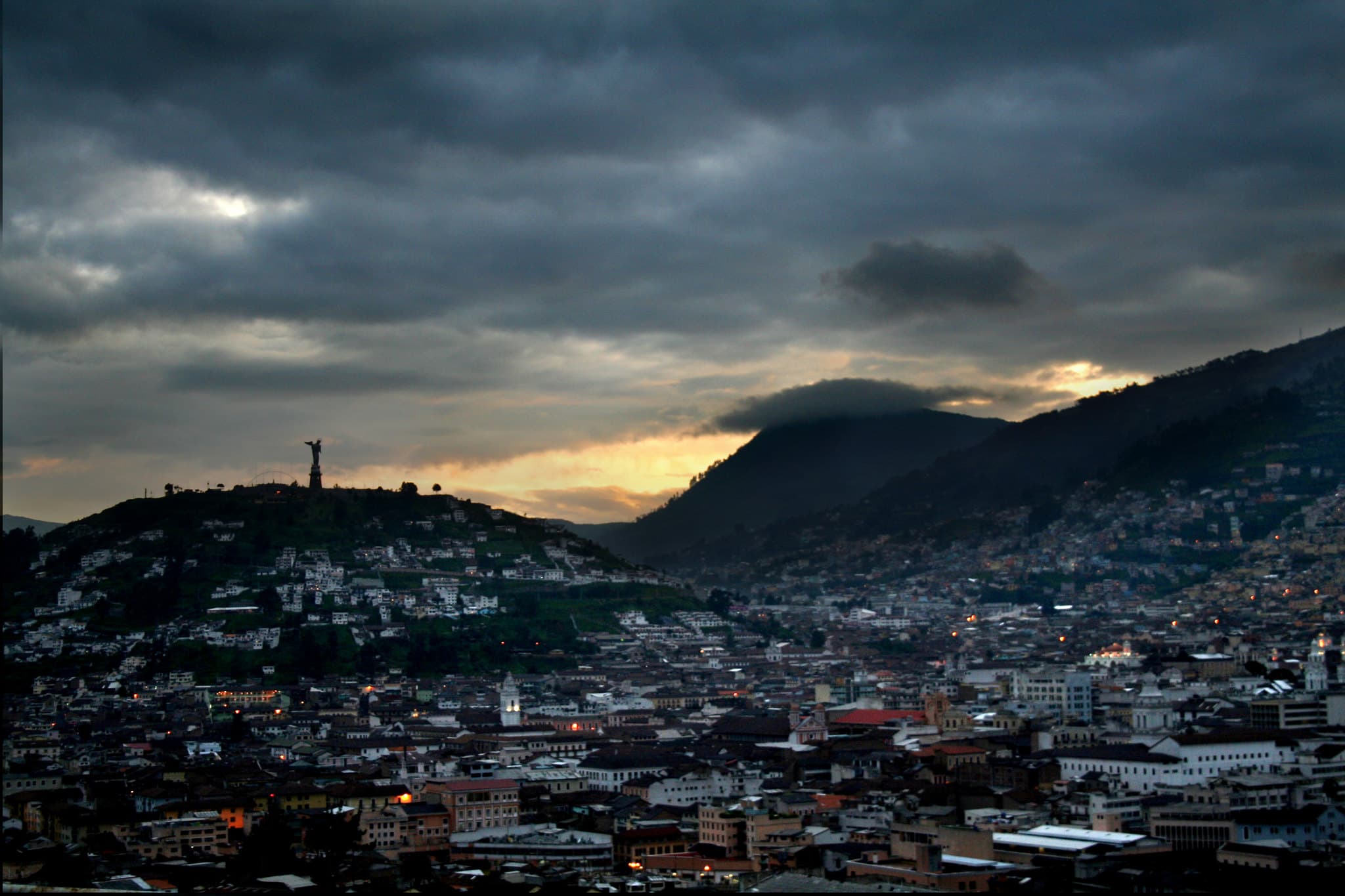 Quito in de avondschemer: de daken van de oude stad onder een donkere wolkenlucht, met links de heuvel El Panecillo met het beeld van de Maagd en rechts de hellingen van de Pichincha.