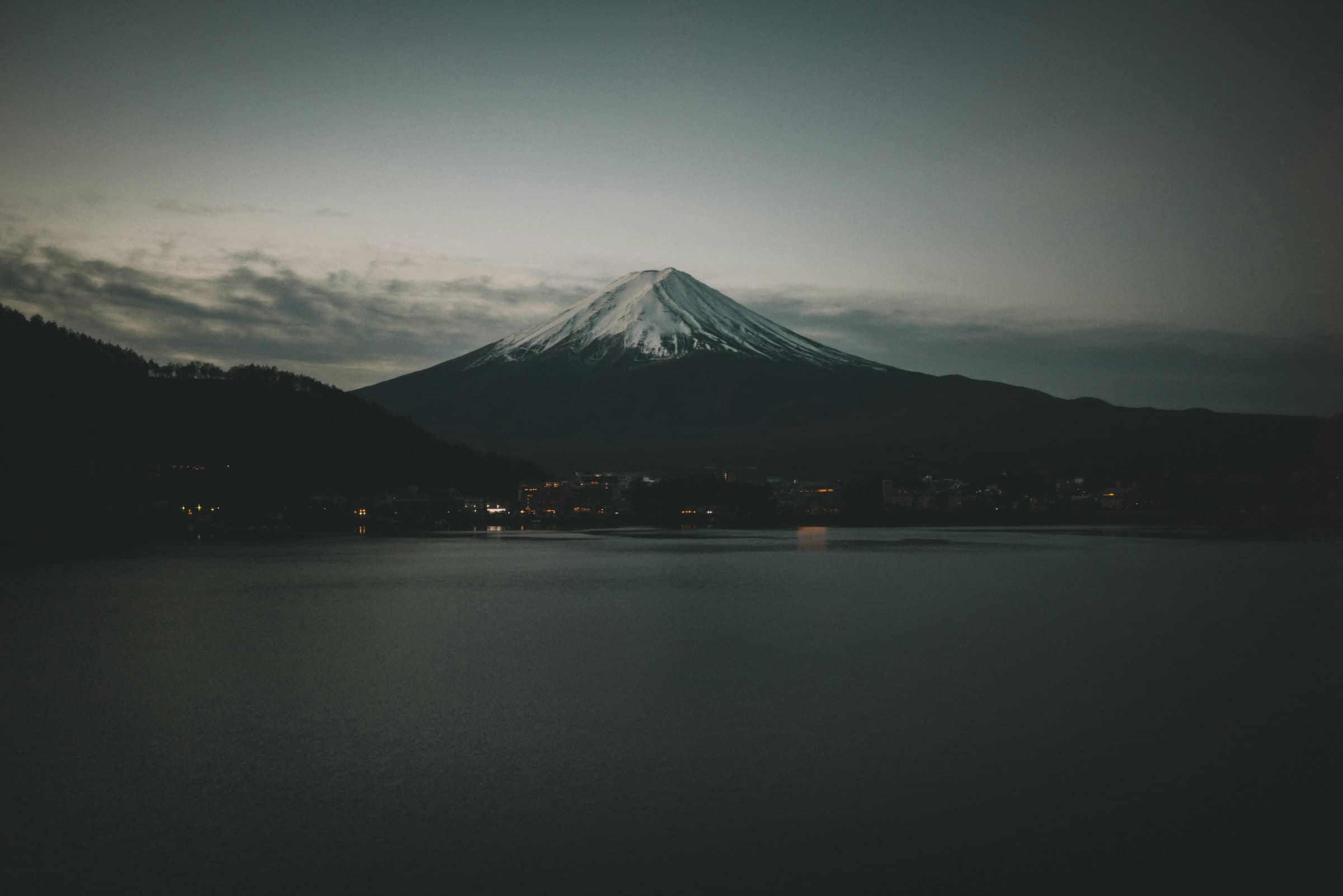 De besneeuwde top van de Fuji in de avondschemering, gezien over het Kawaguchiko-meer, met de lichtjes van het dorp aan de overkant.