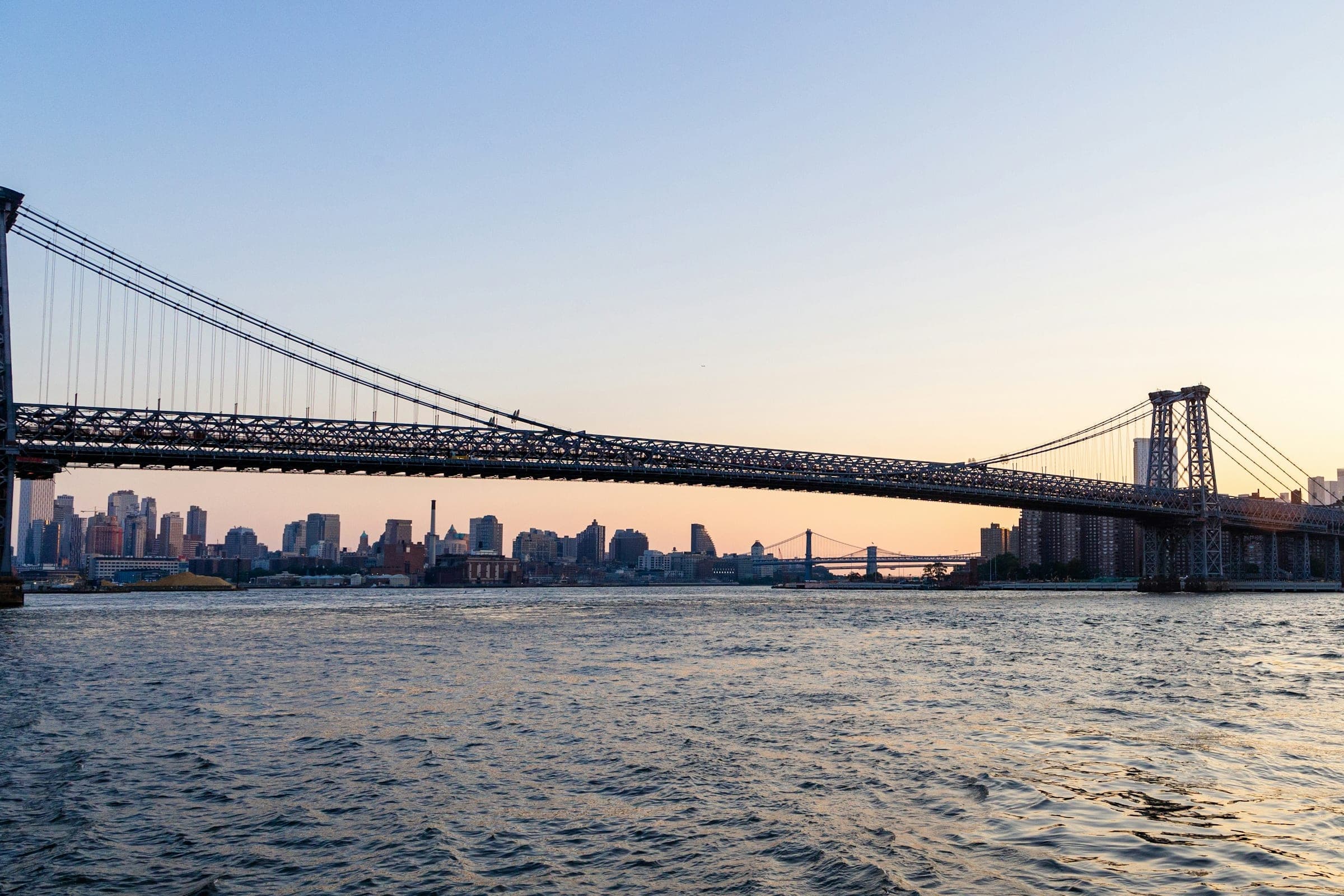 De Williamsburg Bridge over de East River in de schemering, met de skyline van New York erachter.