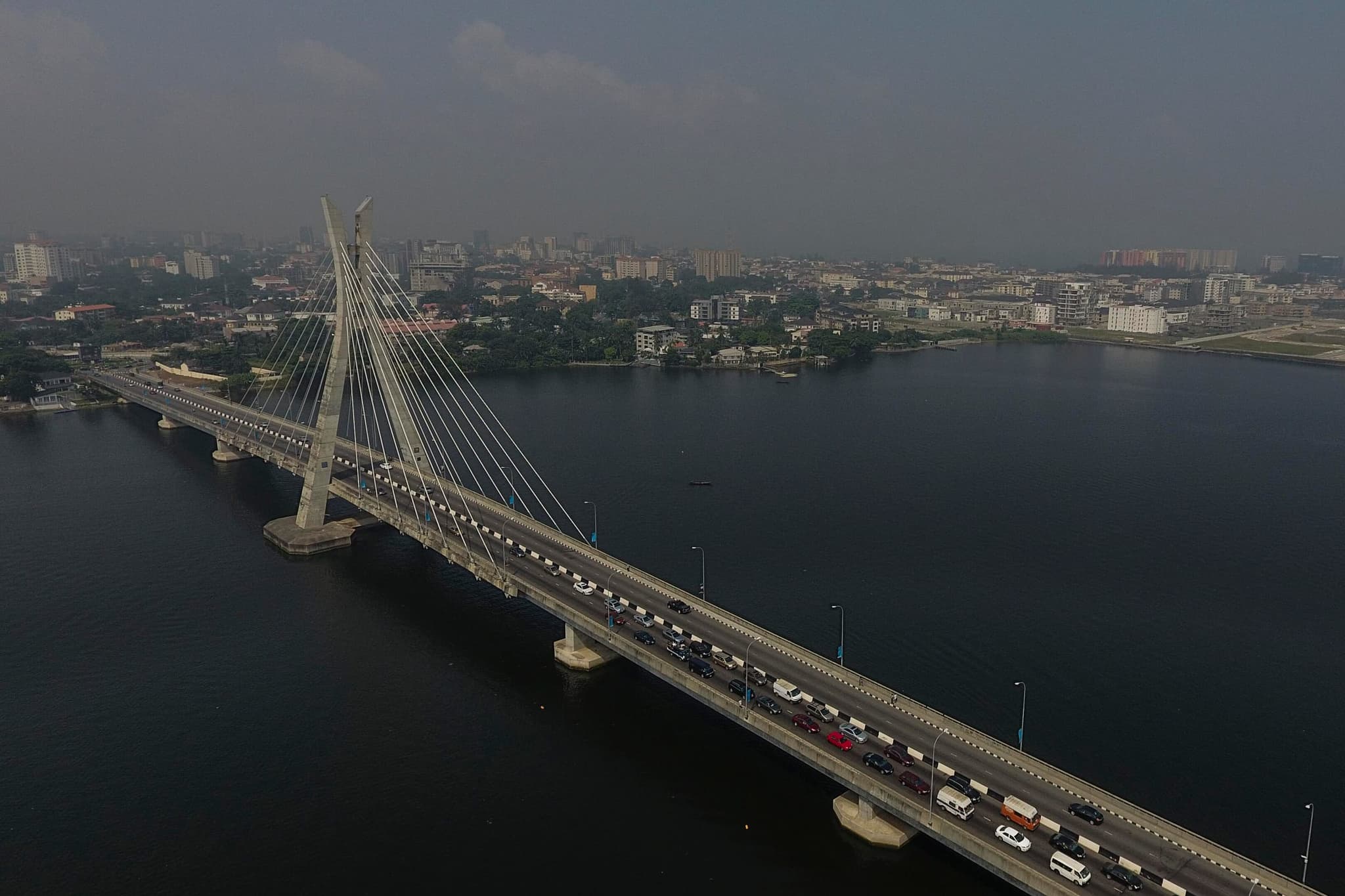 The Lekki-Ikoyi Link Bridge in Lagos from above: the cable-stayed bridge with traffic over the dark lagoon, and the Ikoyi district behind it under a hazy sky.