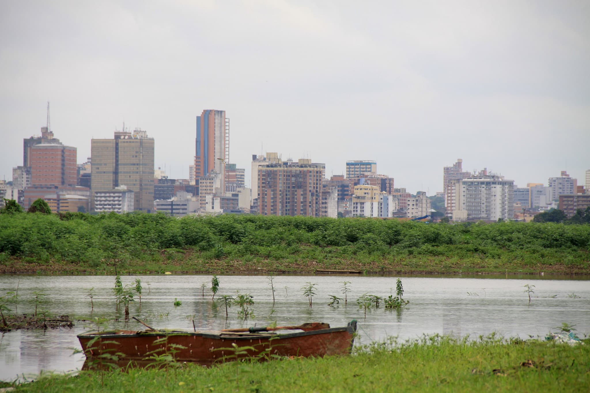 De skyline van Asunción vanaf de oever van de baai: torens en flats achter een groene strook, met op de voorgrond een houten roeiboot op het ondergelopen gras.