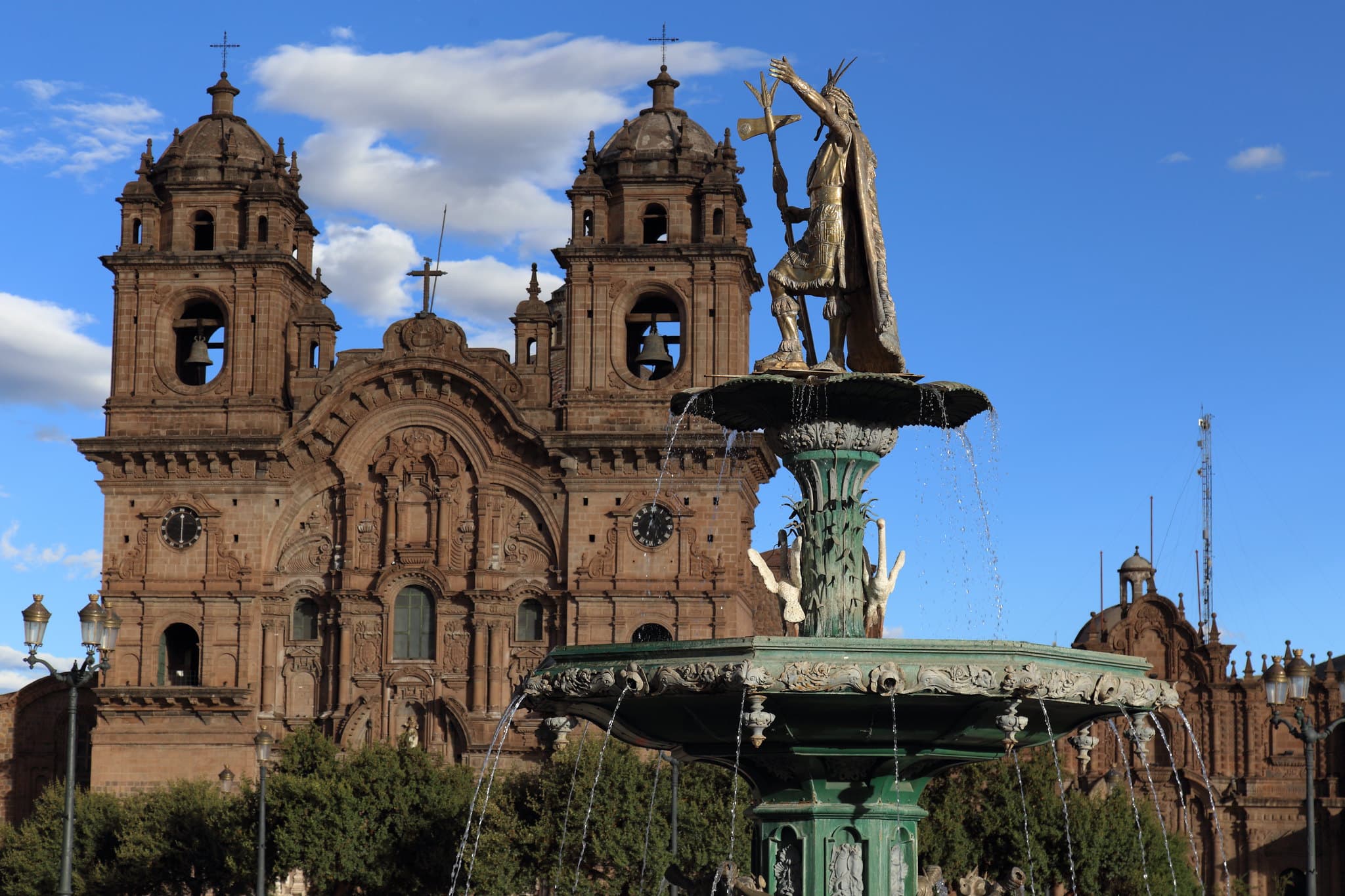 De Plaza de Armas van Cusco onder een blauwe lucht: de fontein met het bronzen beeld van de Inca Pachacútec voor de twee klokkentorens van de Iglesia de la Compañía de Jesús.