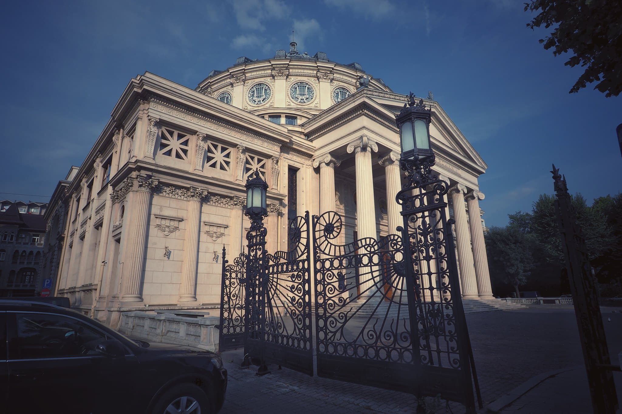 Het Roemeens Atheneum in Boekarest in het avondlicht: de zuilengalerij en de koepel van de ronde concertzaal, gezien door het smeedijzeren hek met de twee lantaarns.