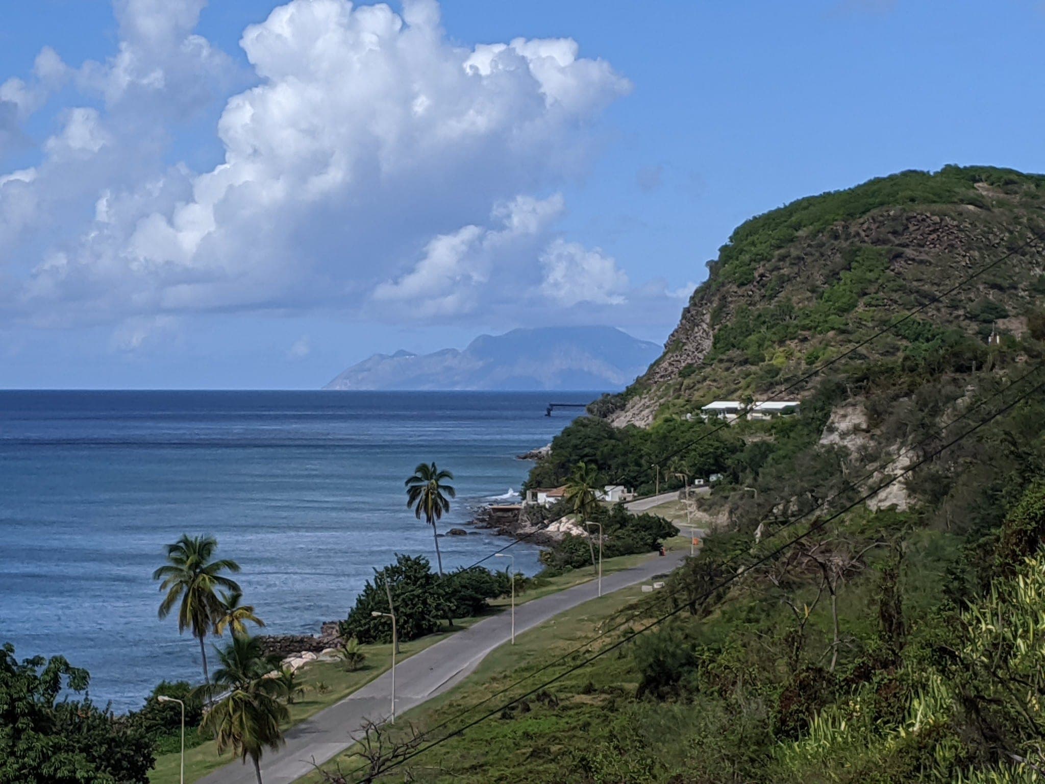 De kustweg langs Oranje Baai bij Oranjestad op Sint-Eustatius, met palmen, de pier en het eiland Saba aan de horizon.