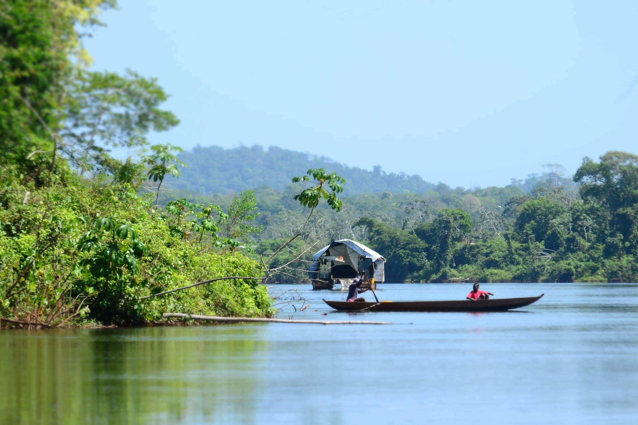 A boat with two people aboard on the Upper Suriname, between densely wooded banks with hills in the mist.