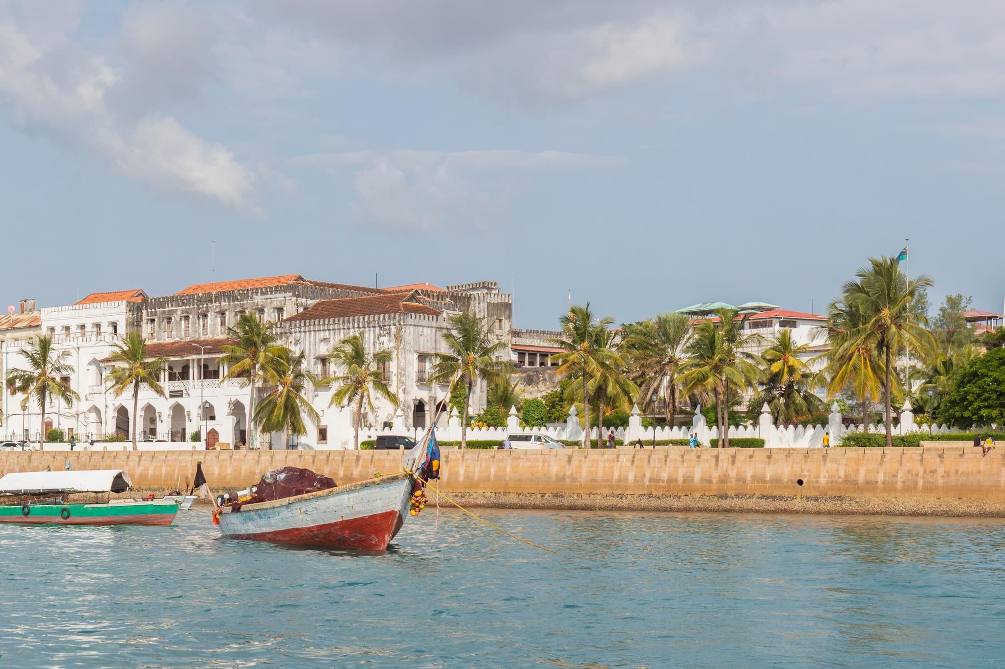The seafront of Stone Town seen from the water: a wooden boat on turquoise sea, behind it a quay wall, white buildings with red roofs and a row of palm trees under a lightly clouded sky.
