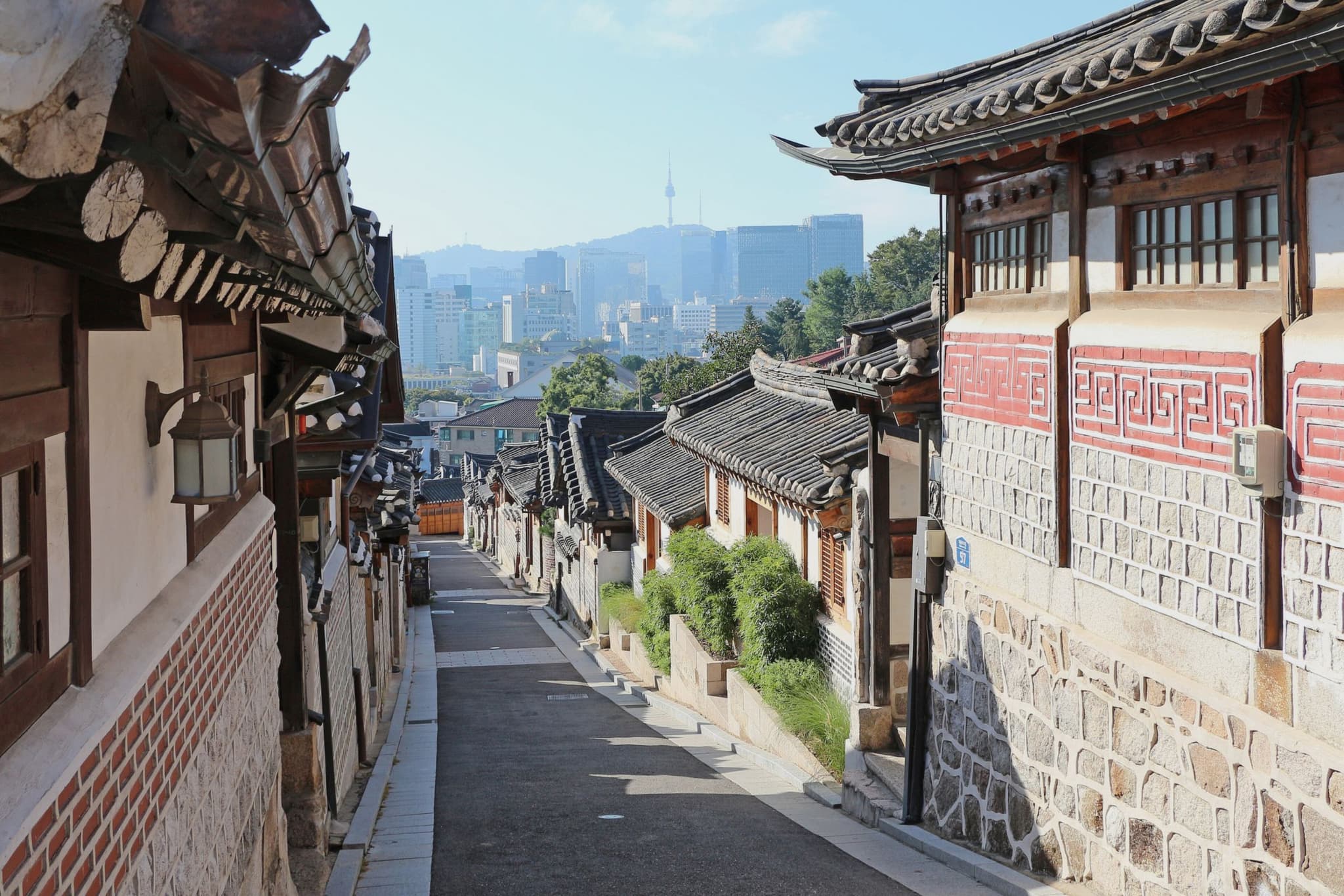 An alley in Bukchon Hanok Village in Seoul: traditional houses with grey tiled roofs and walls of stone and red brick, with the N Seoul Tower above the city in the distance.